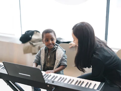 A young child plays an electronic keyboard while an adult sits nearby, smiling and watching attentively.