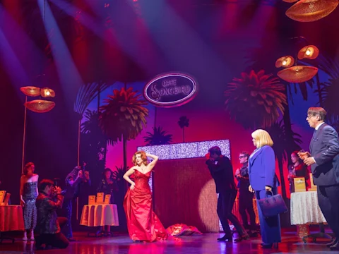 Actors perform on a stage set as "Café Songbird," featuring a woman in a red dress posing while others stand and photograph her under colorful lights and palm tree props.