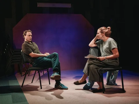 Two people in conversation sit on chairs facing each other in a dimly lit room with a purple backdrop. One leans forward with hand on chin, while the other sits back with arms crossed and an attentive expression.