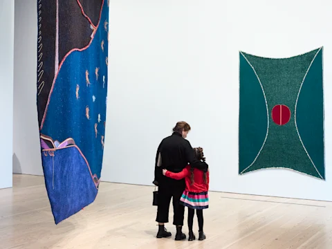 Two people stand closely together in an art gallery, looking at two large colorful textile artworks hanging on white walls.