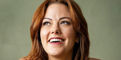 A person with shoulder-length auburn hair smiles while looking upwards, set against a plain, neutral background.