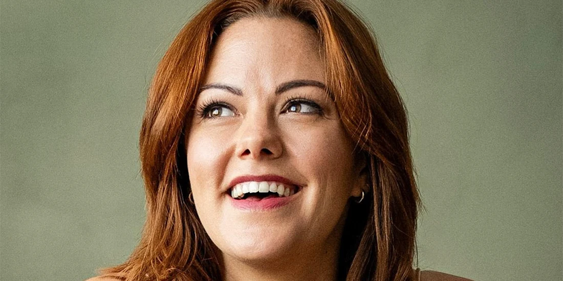 A person with shoulder-length auburn hair smiles while looking upwards, set against a plain, neutral background.