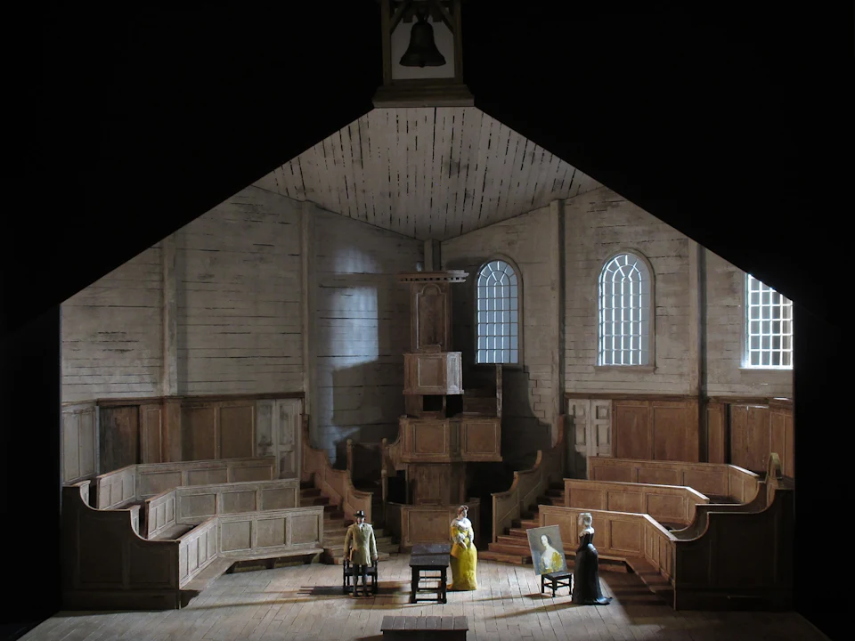 A scale model of a wooden church interior with pews, a central pulpit, and four small human figures positioned on the floor.
