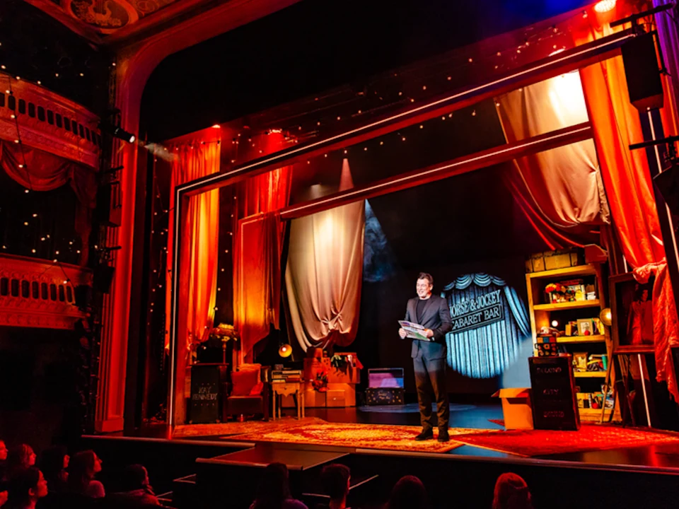 A performer stands holding papers on a brightly lit theater stage decorated with curtains, props, and vintage furniture, facing an audience.