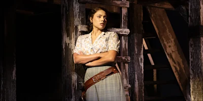 A woman with her arms crossed leans against a wooden ladder in a dimly lit, rustic setting.