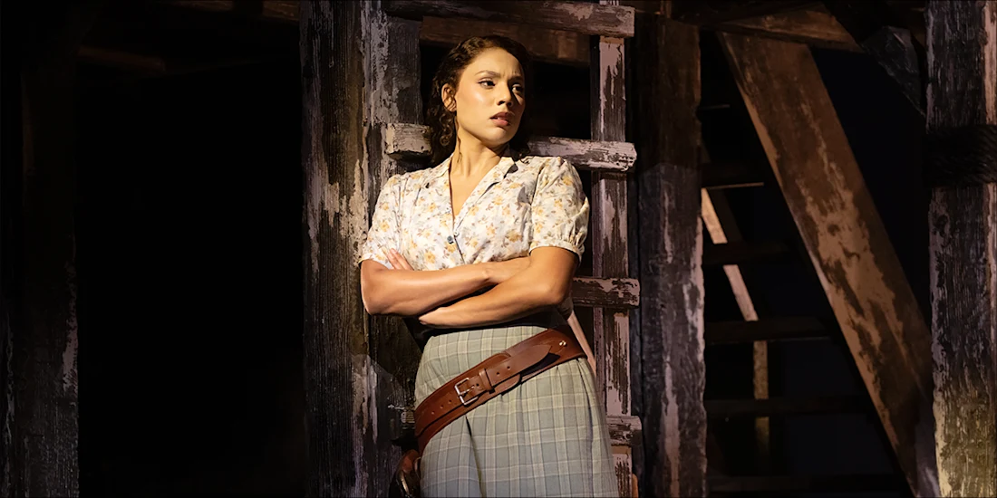 A woman with her arms crossed leans against a wooden ladder in a dimly lit, rustic setting.
