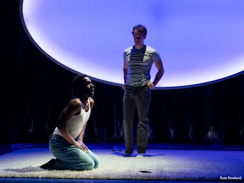 Two men perform on a stage under dramatic blue lighting; one kneels on the floor looking up, while the other stands behind him, watching.