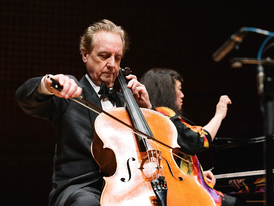 A man in a suit plays the cello with a bow while a woman in colorful clothing plays the piano in the background on stage.
