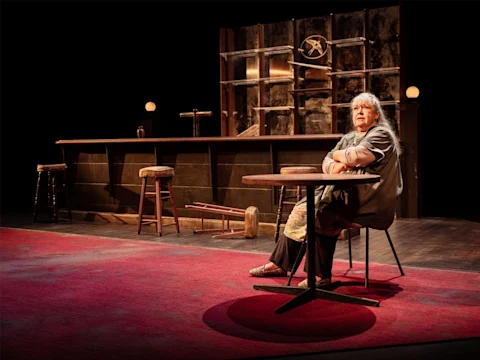 An older woman sits alone at a round table on a red carpeted stage, with empty bar stools and a bar counter in the background.