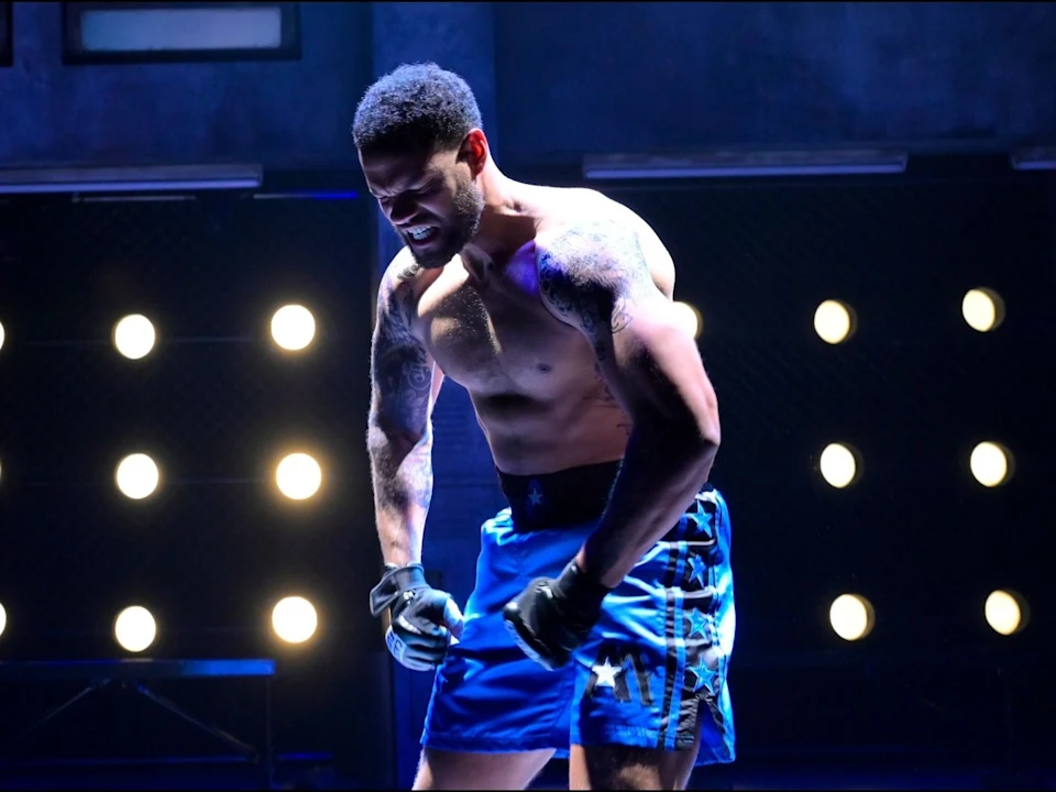 A male MMA fighter in blue shorts and gloves stands under bright lights, flexing his arms and displaying a determined expression.