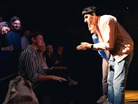 Production photo of It’s a Motherf**king Pleasure in Washington, showing a man in a pink shirt performs energetically in front of a seated audience. Another person stands beside him. The audience members seem engaged and smiling.