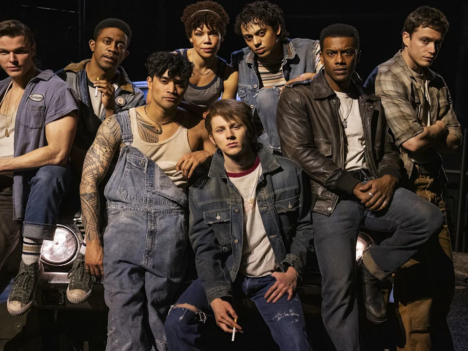 A group of seven young adults in casual clothing pose together in front of a car, some sitting and some standing, in a dimly lit setting.