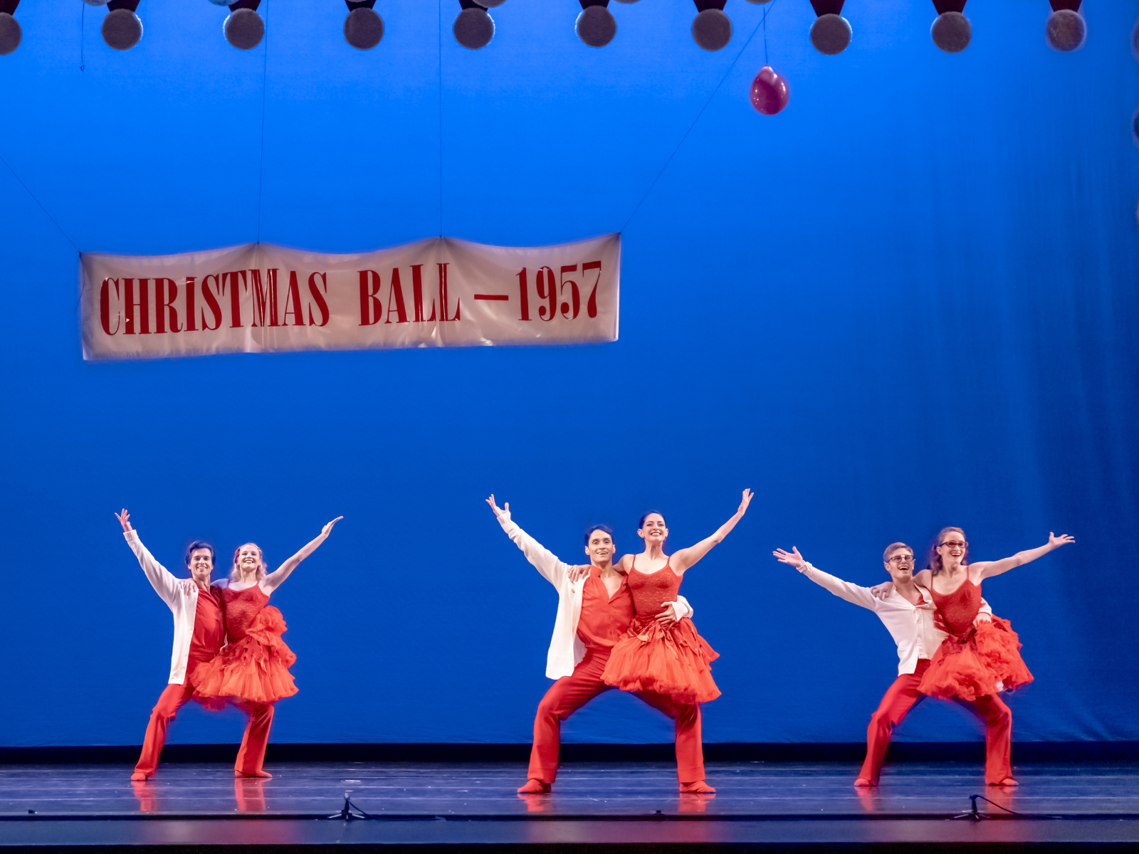 Six dancers in red costumes perform under a banner reading "Christmas Ball - 1957" on a stage with a blue backdrop.
