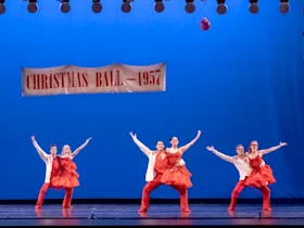 Six dancers in red costumes perform under a banner reading "Christmas Ball - 1957" on a stage with a blue backdrop.