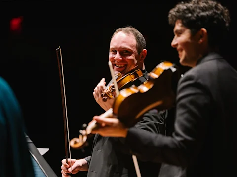Two violinists perform on stage, one smiling and looking at the other, both holding violins and bows under warm stage lighting.