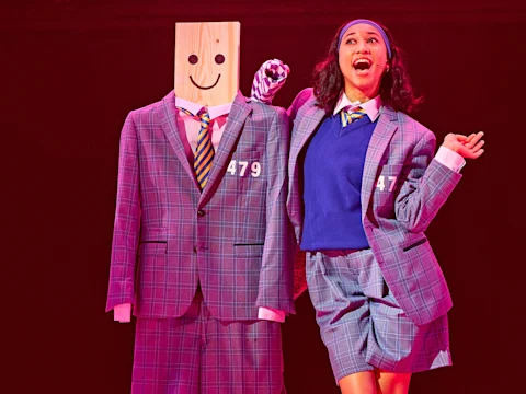 A performer in a plaid suit poses next to a mannequin wearing matching clothes with a wooden block for a head, featuring a drawn-on smiley face, on a brightly lit stage.