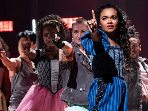 A group of performers on stage, with the woman in front wearing a blue dress and pointing upwards.