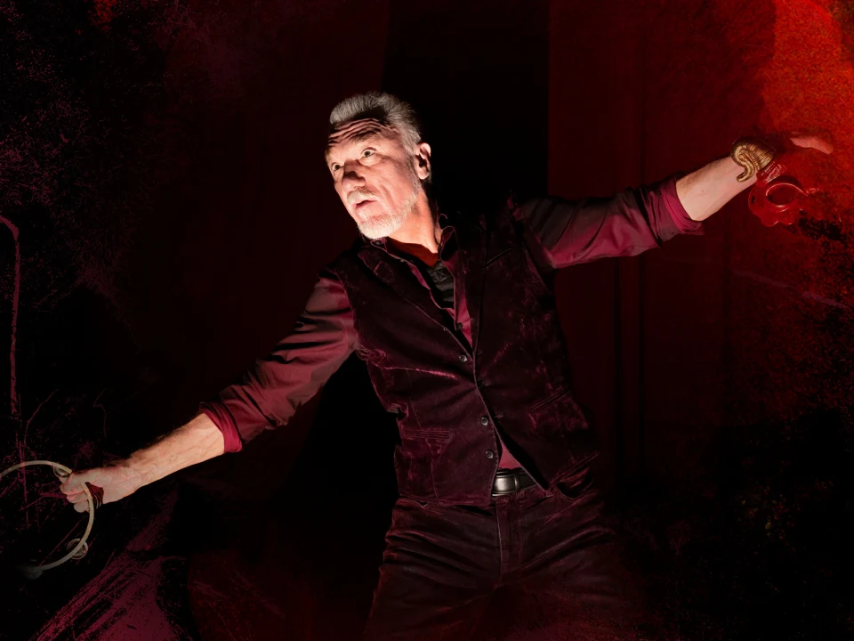 A person in a dark red shirt and vest strikes a dynamic pose against a dramatic red and black background.