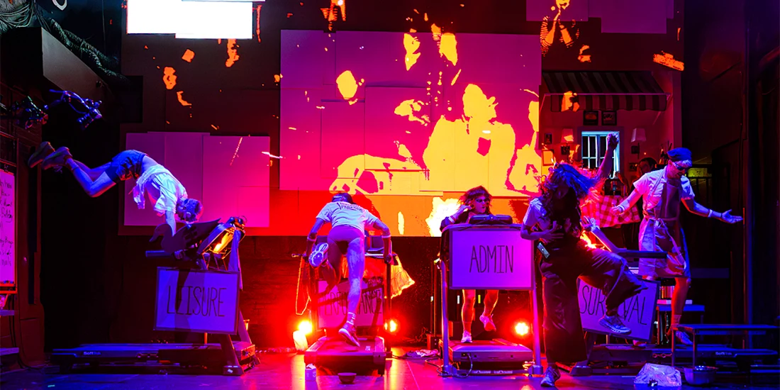 Six performers act energetically on stage with bright red-orange lighting, props labeled "LEISURE," "ADMIN," and "ORGANIC," and abstract visuals projected in the background.