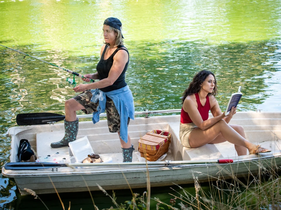 Two people in a boat on a lake: one is fishing while standing, and the other is sitting and reading.