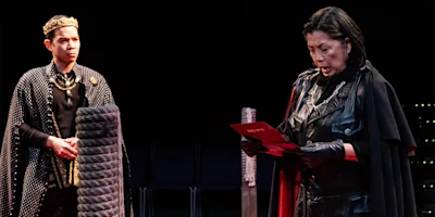 Two actors in medieval costumes perform on stage; one wears a crown and cape, while the other in black reads from a red paper.