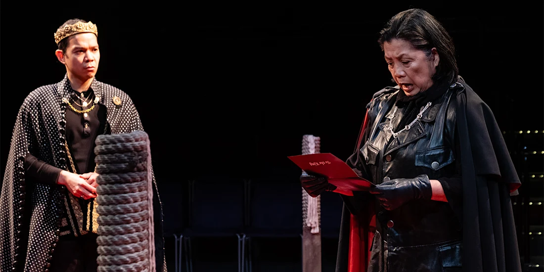Two actors in medieval costumes perform on stage; one wears a crown and cape, while the other in black reads from a red paper.