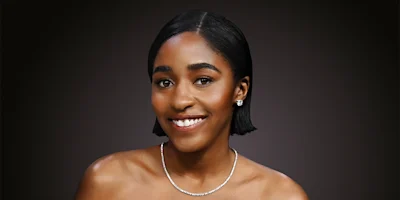 A woman with short, straight black hair, wearing a strapless top, a diamond necklace, and stud earrings, smiles against a dark gradient background.