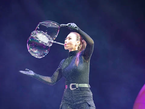 A performer dressed in black creates large bubbles on stage with a bubble wand against a dark background.