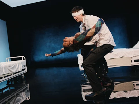 Production photo of Gruesome Playground Injuries in New York, showing a man with a bandaged head holds and dips a woman dramatically in a stage setting with two hospital beds in the background.