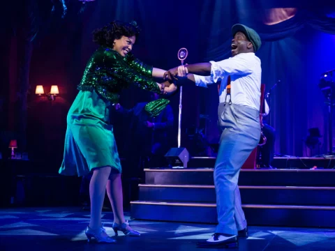 Production photo of Ain't Misbehavin' in Chicago with Alanna Lovely and James T. Lane dancing energetically on stage under blue lighting, with a retro microphone and musicians visible in the background.