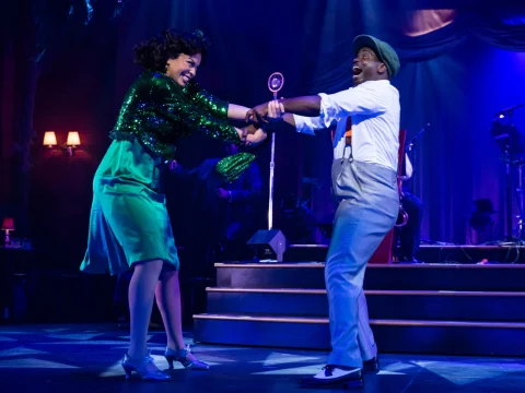 Production photo of Ain't Misbehavin' in Chicago with Alanna Lovely and James T. Lane dancing energetically on stage under blue lighting, with a retro microphone and musicians visible in the background.