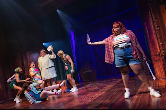 Six performers on stage act out a lively scene, with one person standing in front holding a paper and others reacting energetically in the background.