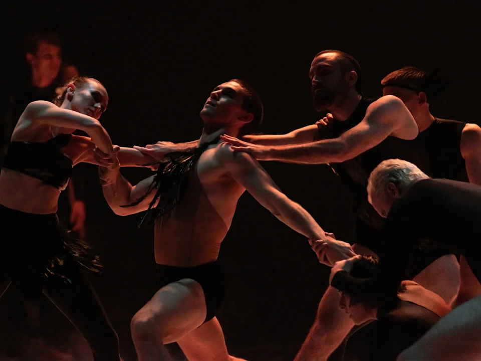 A group of dancers in dark stage lighting perform an expressive routine, reaching and holding each other in dynamic poses.
