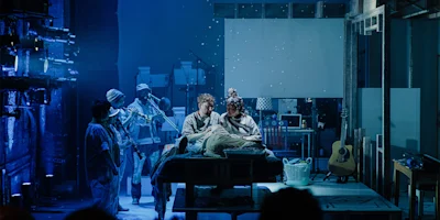 A group of people in costumes stand around a person lying on a table under dramatic blue stage lighting, with various props and set pieces visible in the background.