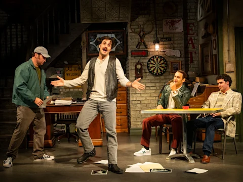 Four men on a stage set resembling a cluttered room; one man stands gesturing dramatically while the others sit or stand, watching him. Papers are scattered on the floor.