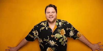 Smiling man with a beard stands against a bright yellow wall, wearing a black floral shirt with yellow and green patterns.