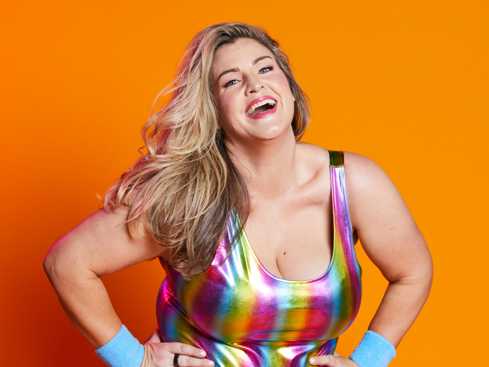 Smiling woman with long blonde hair wears a shiny rainbow tank top and blue wristbands, standing against an orange background.