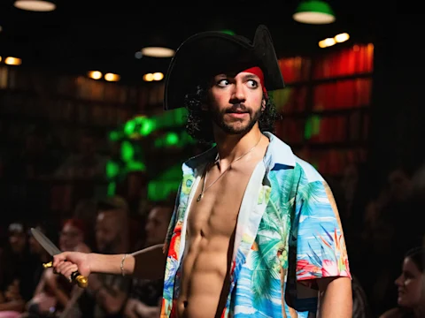 A man in a tropical shirt and pirate hat holds a dagger, standing in a dimly lit room with people seated in the background.