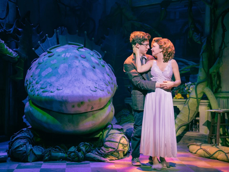 Two actors (Andrew Barth Feldman and Sarah Hyland) in a dramatic embrace on stage, with a large, plant-like prop in the background.