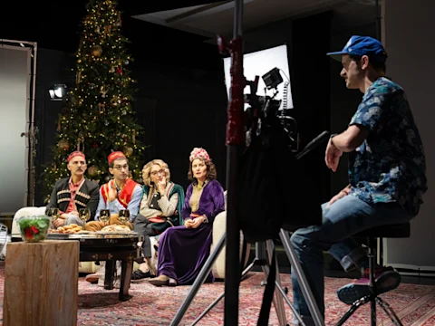 A group of four people in colorful costumes sit by a table with food, facing a camera operator in a studio with a decorated Christmas tree in the background.