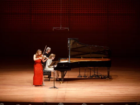 Two musicians perform on stage: one playing the violin in a red dress, and the other playing a grand piano. The stage has a wooden floor and a perforated backdrop. An audience is partially visible.