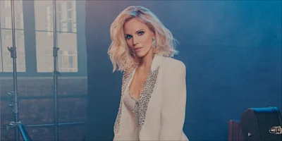 A woman with blonde hair wearing a white blazer with crystal lapels poses in front of a blue backdrop.