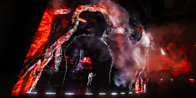 A performer stands on stage surrounded by large, illuminated, tree-like structures with red and orange lighting and smoke effects.