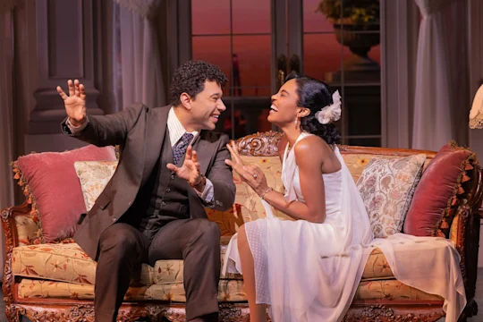 A man in a suit and a woman in a white dress sit on a decorative sofa, smiling and gesturing enthusiastically to each other in an elegant room.