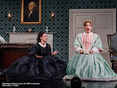 Two actors in period costumes perform on stage in front of a patterned wall and a painting of George Washington; one appears surprised, the other gestures while seated on the floor.