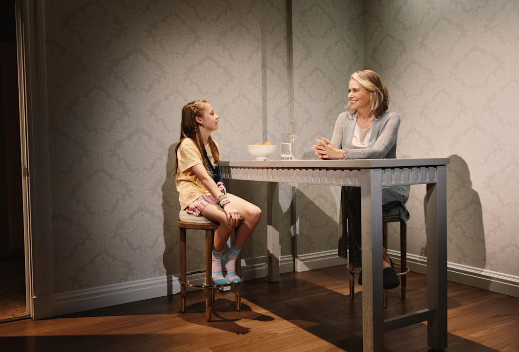 A young girl and a woman sit across from each other at a tall table, conversing in a simply decorated room with patterned wallpaper and wooden floors.