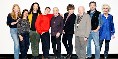 Nine adults stand in a row against a white background, posing for a group photo. They are dressed in casual clothing and are smiling.