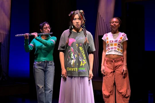 Three women are on stage; one plays a flute, the center woman sings or speaks, and the third stands behind them. Stage lighting is blue and purple.