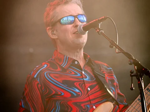 A musician wearing sunglasses and a red, blue, and black swirled shirt sings into a microphone while playing guitar on stage.