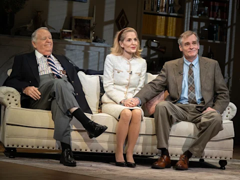 Three adults in formal attire sit on a beige sofa in a well-lit living room. The man and woman on the right are holding hands.